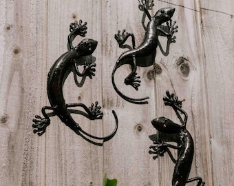 May include: Three black metal lizard sculptures are mounted on a light-colored wooden fence. The lizards have detailed feet and tails, with their bodies curved in various poses. The fence has visible wood grain.