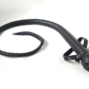 Puede incluir: Látigo de cuero negro con un mango trenzado y una correa de muñeca en bucle. El cuerpo del látigo también está trenzado, afinándose hacia un extremo delgado con varios hilos de cuero. El mango tiene un diseño redondeado y texturizado.