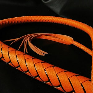 Puede incluir: Un l&aacute;tigo de cuero trenzado con un borla de cuero marr&oacute;n. El l&aacute;tigo es naranja y tiene un dise&ntilde;o trenzado negro.