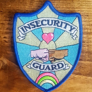 Puede incluir: Un parche bordado azul y beige con el texto "Insecurity Guard" en una pancarta. Dos puños, uno rosa y otro marrón, se golpean. Un corazón rosa y un arcoíris también están bordados en el parche.