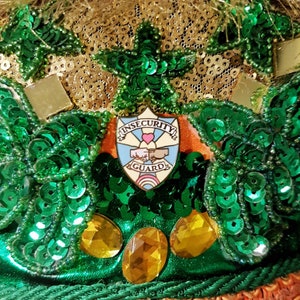 May include: A gold and green pin with the words "Insecurity Guard" and a heart with a fist on top of it. The pin is on a green and gold background with sequins.