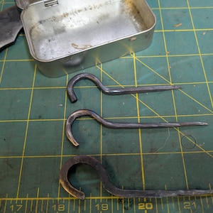 Pode incluir: Dois ganchos de metal com uma extremidade arredondada e uma extremidade pontiaguda. Os ganchos estão em um tapete de corte verde com uma régua.