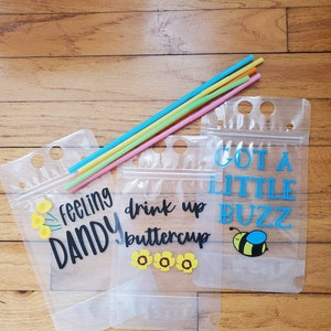 May include: Clear plastic drink pouches with colorful straws. The pouches have text that reads "feeling DANDY", "drink up buttercup", and "GOT A LITTLE BUZZ" with flower and bee graphics. The straws are pink, yellow, green, and blue.
