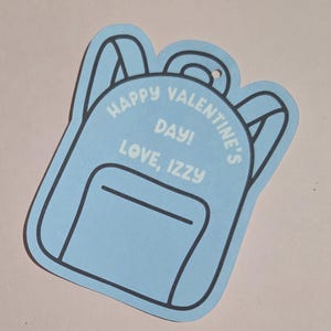 May include: A light blue backpack-shaped Valentine's Day card with black outlines. The card features the text "HAPPY VALENTINE'S DAY! LOVE, IZZY" in white. The card has a small hole at the top for hanging.
