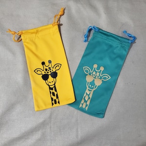 May include: Two drawstring eyeglass pouches. One is yellow with a black giraffe wearing sunglasses. The other is teal with a gold giraffe wearing sunglasses. Both pouches have a drawstring closure.