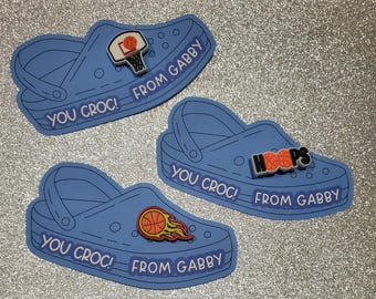 May include: Three blue Croc shoe-shaped cutouts with basketball-themed charms. Each cutout has the text "YOU CROC! FROM GABBY" in white. One charm shows a basketball and hoop, another says "HOOPS", and the third has a basketball with flames.