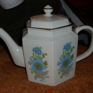 May include: White ceramic teapot with a floral design featuring blue flowers and gold trim. The teapot has a hexagonal shape with a lid and a curved spout. A classic piece for serving tea.