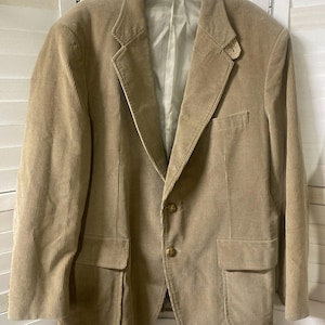 May include: A tan corduroy blazer with a notched lapel, two front pockets, and two-button closure. The jacket is hanging on a clear hanger against a white shutter background. The blazer has a classic, tailored design.