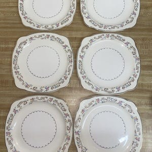 Può includere: Set di sei piatti da portata quadrati con un motivo floreale e un bordo dorato. I piatti sono bianchi con un centro color crema e un bordo smerlato.