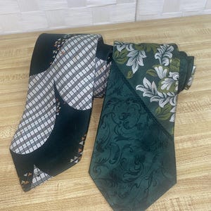May include: Two neckties. One is black with a white and black chequered pattern and orange accents. The other is dark green with a floral design in white and green. Both are folded on a wooden surface.