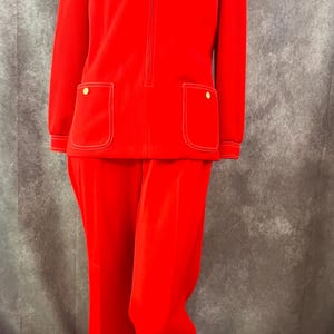 Pode incluir: Um fato vintage de veludo cotelê vermelho brilhante. O casaco tem fecho de correr, dois bolsos frontais com botões dourados e costuras brancas. As calças a condizer são largas. O fato é exibido num manequim.