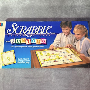 Pode incluir: Uma caixa de Scrabble Crossword Game para Juniors com um design azul e branco. A caixa apresenta o tabuleiro do jogo e duas crianças a jogar. O texto na caixa diz "O jogo de palavras 'perfeito' para crianças!"