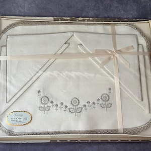 May include: A vintage place mat set in a box. The set includes a large rectangular place mat, two smaller mats, and a napkin, all in white with gray floral embroidery and a gray lace trim. A pink ribbon is tied around the set.