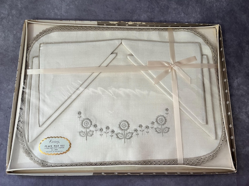 May include: A vintage place mat set in a box. The set includes a large rectangular place mat, two smaller mats, and a napkin, all in white with gray floral embroidery and a gray lace trim. A pink ribbon is tied around the set.