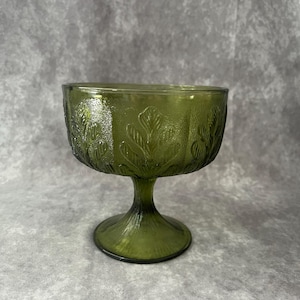 May include: A green glass pedestal bowl with a leaf design. The bowl has a wide, round base and a tall, slender stem.