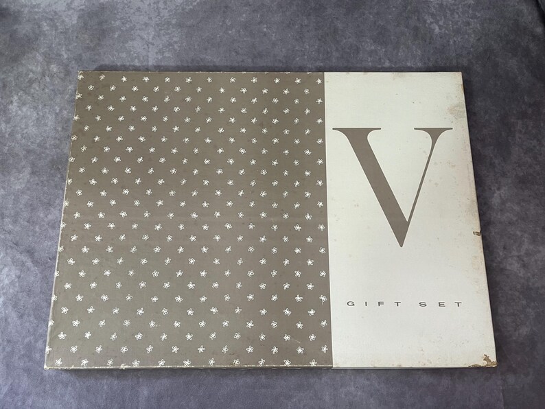May include: Rectangular gift set box with a two-tone design. The left side features a gray background with white star-shaped patterns. The right side is off-white with a large gray letter "V" and the words "GIFT SET" in a serif font.