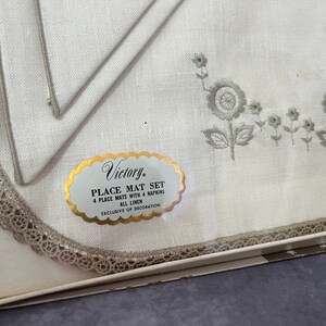 May include: A white linen place mat set with four mats and four napkins. The mats and napkins have a floral embroidery design in gray. The set includes a label that reads "Victory Place Mat Set".