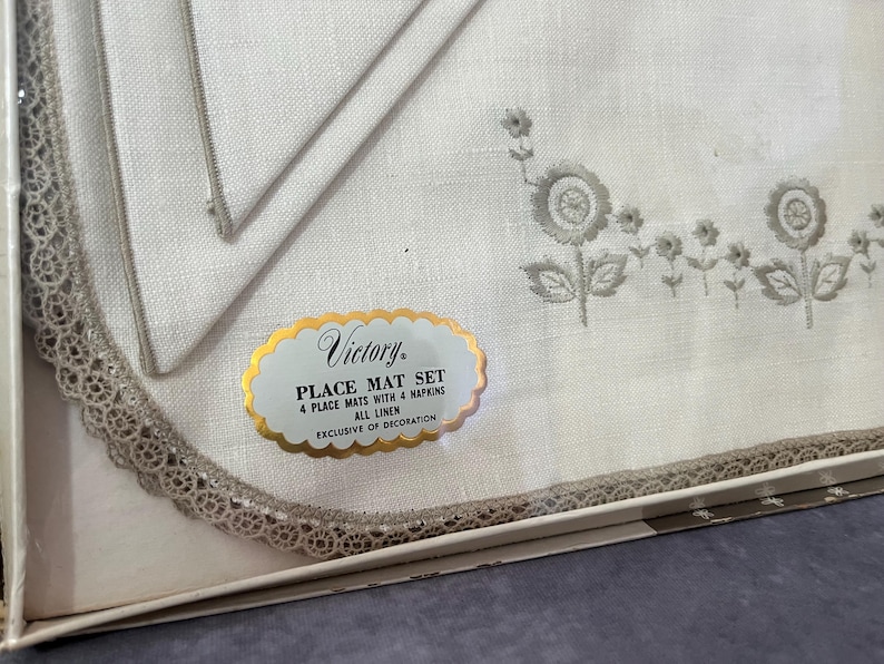 May include: A white linen place mat set with four mats and four napkins. The mats and napkins have a floral embroidery design in gray. The set includes a label that reads "Victory Place Mat Set".