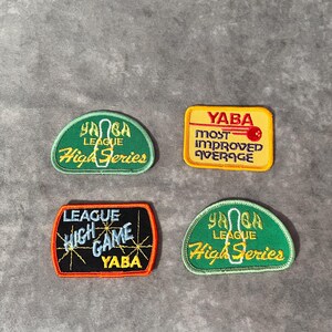 May include: Four embroidered patches with text. Two are green with "HANGA LEAGUE High Series" in yellow. One is yellow with "YABA MOST IMPROVED AVERAGE" in red. The last is black with "LEAGUE HIGH GAME YABA" in white and yellow.