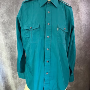 Puede incluir: Camisa abotonada de color verde azulado con dos bolsillos en el pecho y charreteras. La camisa de manga larga tiene un cuello clásico y puños abotonados. La camisa está hecha de una tela de color sólido y tiene un corte entallado.