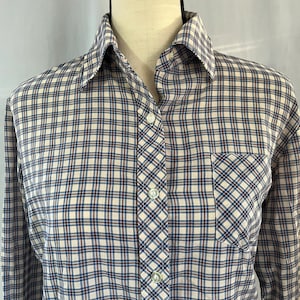 Puede incluir: Una camisa de manga larga con botones, fondo blanco y estampado de cuadros azul, marrón y blanco. La camisa tiene un solo bolsillo en el pecho.