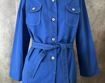 May include: A long-sleeved, blue button-down shirt with white stitching and a matching belt. The shirt has two large pockets on the front and a collar.