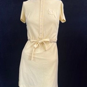 May include: Pale yellow dress with short sleeves and a high collar. The dress features a button-up front, a small pocket on the chest, and a matching tie belt at the waist. The dress is displayed on a mannequin against a dark background.