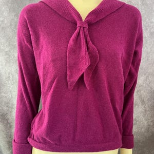 May include: A magenta knitted sweater with a sailor-style collar and a bow tie. The sweater has long sleeves and a ribbed texture.