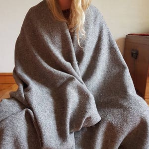Może przedstawiać: Szary wełniany koc z frędzlami na krawędziach. Koc jest zarzucony na osobę siedzącą na drewnianej podłodze.