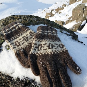 Puede incluir: Un par de mitones tejidos en marrón y crema con un patrón geométrico, descansando sobre una roca cubierta de nieve. Los mitones están hechos de un material grueso y cálido, ideal para el clima frío. El fondo muestra un paisaje nevado con rocas.