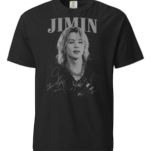 Puede incluir: Camiseta negra con una imagen en escala de grises de una persona con cabello largo y un collar de cadena. La palabra "JIMIN" está impresa encima de la imagen en gris, con un efecto desgastado. Una firma está debajo.