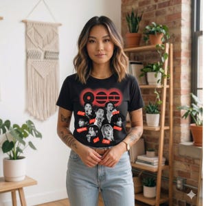 Op de afbeelding: Zwart T-shirt met een rode afbeelding met het woord "000" en afbeeldingen van verschillende mensen. Het shirt heeft korte mouwen en is gemaakt van een zacht materiaal. Het model draagt een blauwe spijkerbroek.