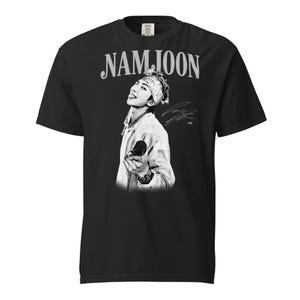 May include: Black t-shirt featuring a black and white image of a person with the name "NAMJOON" in a stylized font above the image. The person is wearing a white jacket and a bandana. A signature is visible on the right side of the image.