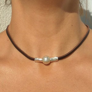 Könnte beinhalten: Eine braune Lederkette mit einer silbernen Perle und einem silbernen Balkenakzent.