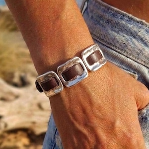 Op de afbeelding: Een armband met drie rechthoekige, gehamerde zilverkleurige metalen schakels verbonden door donkerbruine lederen segmenten. De schakels hebben een gepolijste, onregelmatige textuur en de lederen stukken zijn glad en donker voor een mooi contrast.