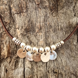 May include: A brown leather necklace with silver beads and round silver charms. The necklace is on a wooden background.