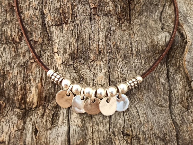 May include: A brown leather necklace with silver beads and round silver charms. The necklace is on a wooden background.