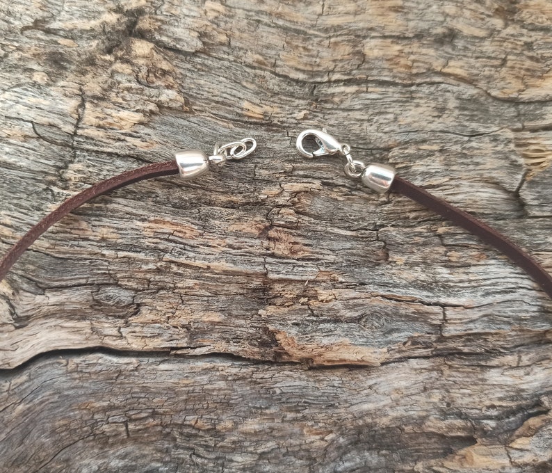 May include: A brown leather cord necklace with silver clasps. The cord is lying on a piece of weathered wood.