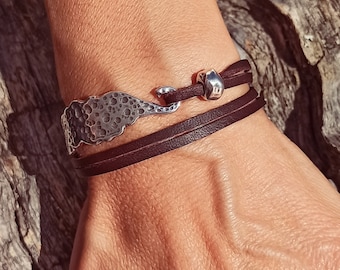 Puede incluir: Un brazalete de cuero marrón con un dije de plata que presenta un diseño abstracto texturizado. El dije tiene un detalle en forma de gancho.