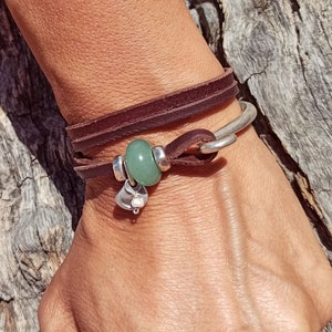 Könnte beinhalten: Ein braunes Lederarmband mit einem silbernen Anhänger und einer grünen Steinperle.
