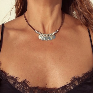 May include: A brown leather choker necklace with silver round charms. The necklace is worn by a person with brown hair.