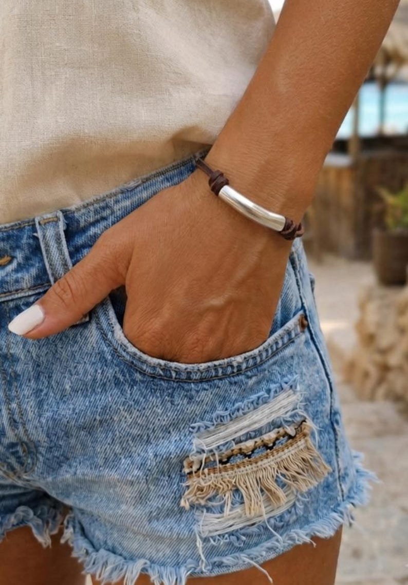 May include: A bracelet featuring a smooth, curved silver-toned metal tube centred on a dark brown leather cord. The cord is knotted on both sides of the metal piece, creating a simple, adjustable design that rests against the wrist.