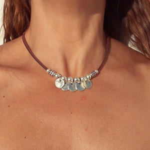 May include: A brown leather choker necklace with silver round charms. The necklace is worn by a person with brown hair.