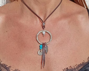 May include: A brown leather cord necklace featuring a silver-toned hammered circular pendant. The pendant is adorned with a small white bead, a bright blue stone, a round silver disc, a silver feather charm, and two thin leather strips.