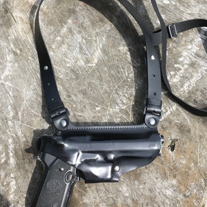 May include: A black leather holster with a shoulder strap for carrying a handgun. The holster is designed to be worn across the body.