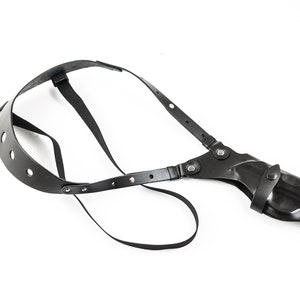 Op de afbeelding: Zwart leren schouderholster met verstelbare banden en een drukknoop sluiting. Het holster is ontworpen om een ​​vuurwapen te dragen.