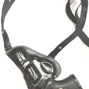 Könnte beinhalten: Ein schwarzes Schulterholster aus Leder mit einer geformten Tasche für einen Revolver. Das Holster ist an verstellbaren Lederriemen mit Metallnieten und Schnallen befestigt, die einen sicheren Halt der Schusswaffe in vertikaler Position gewährleisten.