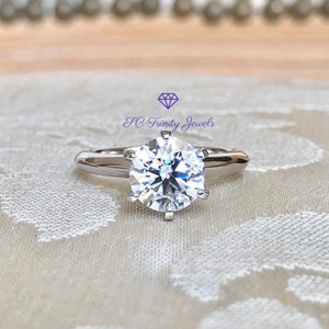May include: A silver solitaire engagement ring with a large, round, clear diamond. The ring is set on a white fabric background.