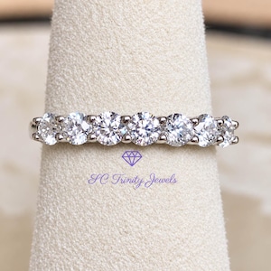 May include: A silver-toned band ring featuring seven round, clear, faceted stones set in a row. Below the ring, the text SC Trinity Jewels is printed in purple, accompanied by a small purple diamond-shaped icon above the brand name.