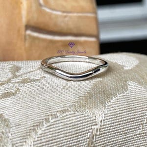 May include: A polished silver-toned curved band ring featuring a smooth, continuous surface. The ring has a distinct wave-like contour along its top edge. A small purple logo with a diamond icon and the text SC Trinity Jewels is visible.